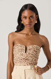 A woman is wearing a strapless top with a leopard print pattern and a center slit detail. She pairs it with high-waisted, light-colored pants. She accessorizes with large gold hoop earrings, a gold bracelet, and multiple gold rings.